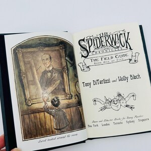 Puede incluir: Un libro abierto con ilustraciones de "The Spiderwick Chronicles: The Field Guide". La p&aacute;gina izquierda muestra a un ni&ntilde;o mirando una pintura. La p&aacute;gina derecha muestra el t&iacute;tulo, los nombres de los autores y la informaci&oacute;n del editor.