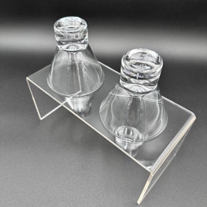 Peut inclure: Deux verres &agrave; shot en verre transparent avec une base large et un sommet &eacute;troit, pos&eacute;s sur un support en acrylique transparent.