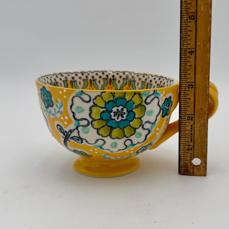 May include: Yellow teacup with a floral design in teal, green, and white. The teacup has a handle and a pedestal base. The teacup is approximately 3 inches tall.