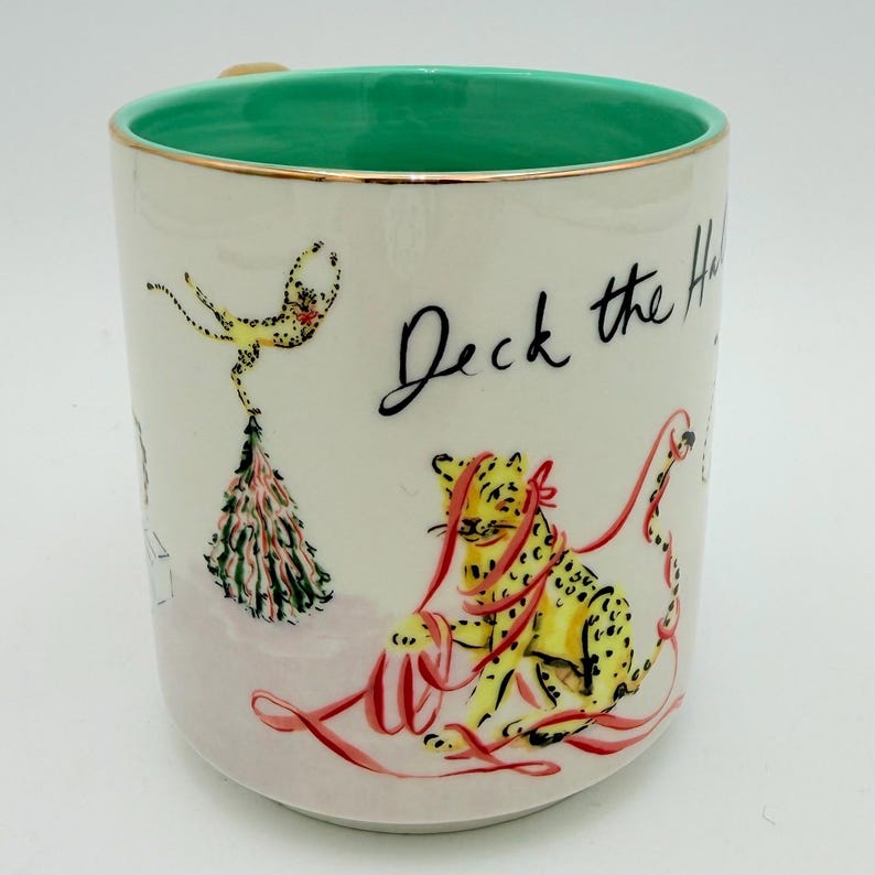 K&ouml;nnte beinhalten: Wei&szlig;e Keramiktasse mit gr&uuml;nem Innenraum und Goldrand. Die Tasse zeigt ein handgezeichnetes Design eines gelben Leoparden mit rotem Band und den Worten "Deck the Halls" in schwarzer Schrift. Ein festliches Urlaubsthema.