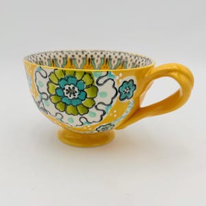 May include: Yellow ceramic teacup with a large floral design in teal, green, and white. The cup has a rounded base, a curved handle, and a decorative rim. The teacup is a bright, cheerful color.