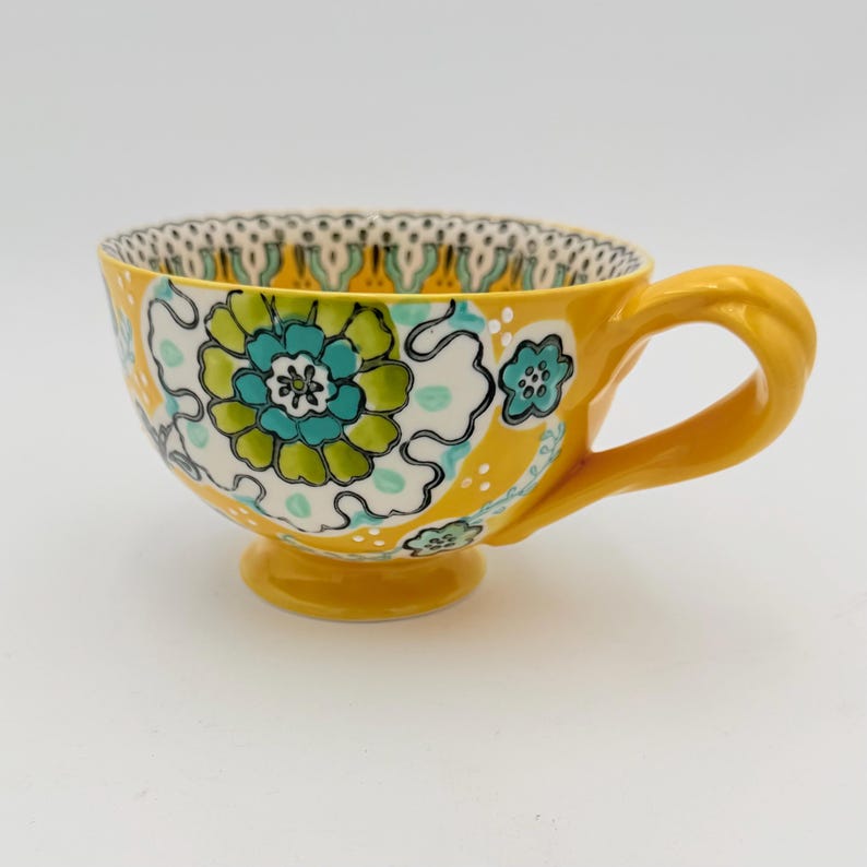 May include: Yellow ceramic teacup with a large floral design in teal, green, and white. The cup has a rounded base, a curved handle, and a decorative rim. The teacup is a bright, cheerful color.
