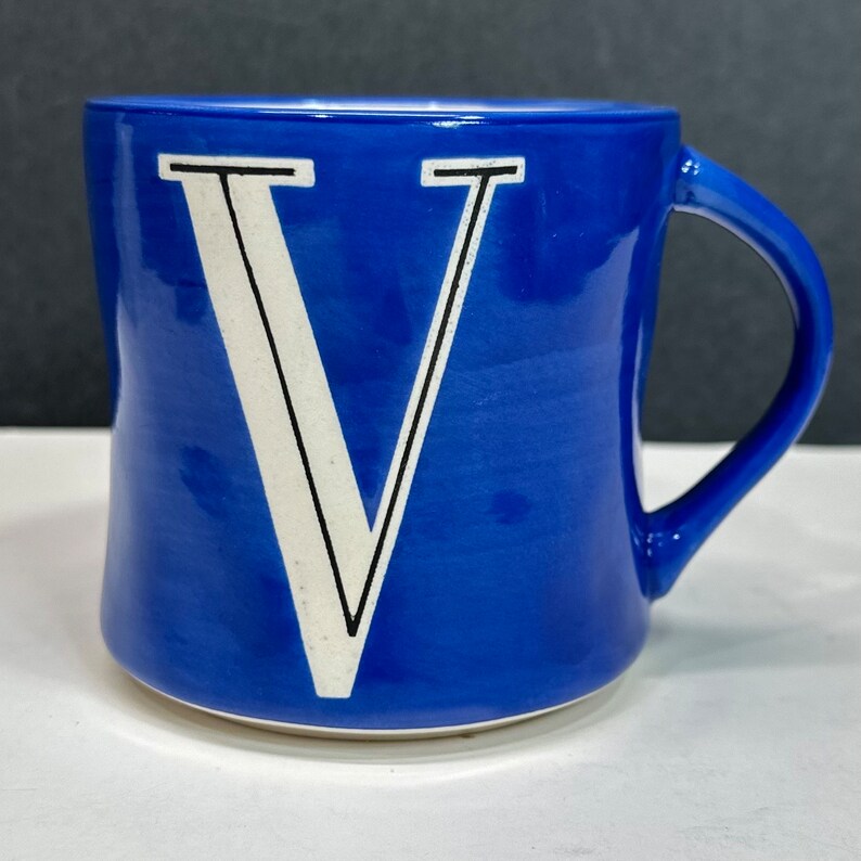 May include: A cobalt blue ceramic mug with a white letter "V" in a bold, sans-serif font. The mug has a curved handle and a slightly flared shape. The letter "V" is outlined in black.