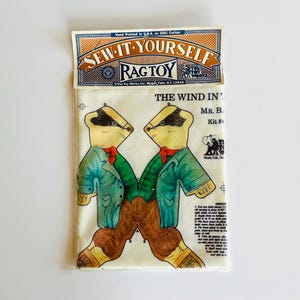 May include: A vintage "Sew-It-Yourself Ragtoy" kit featuring a badger design. The kit includes a printed fabric with two badger illustrations wearing green jackets, red bow ties, and brown pants. The packaging includes text that reads "The Wind In... Mr. B, Kit #4."