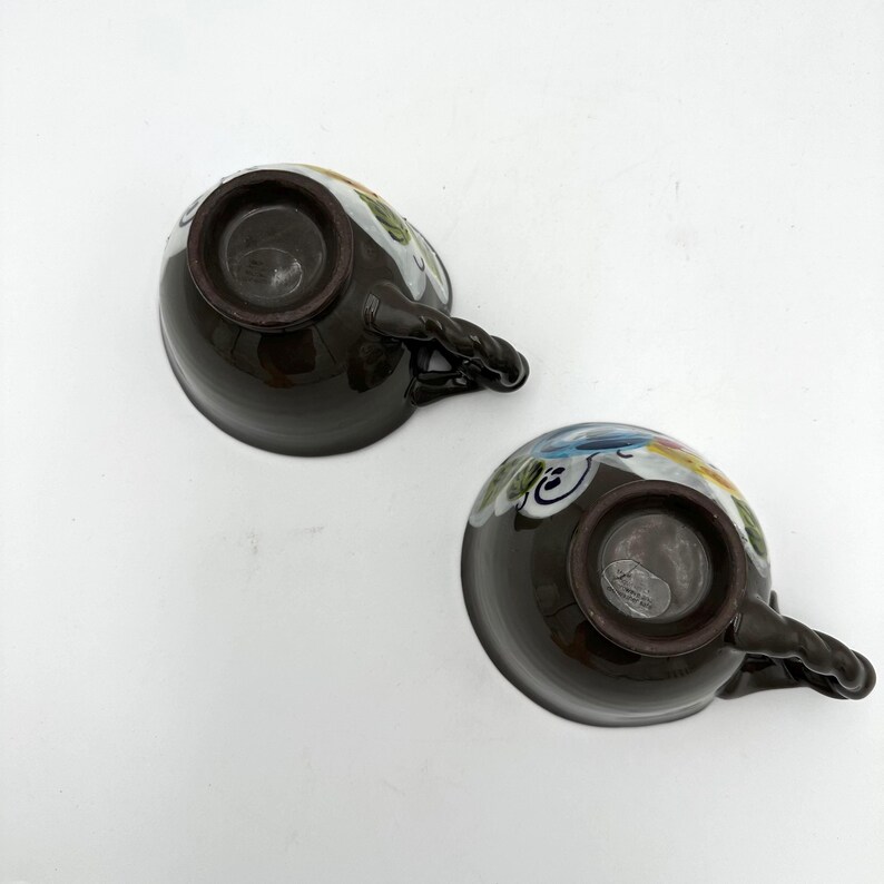 Puede incluir: Dos tazas de t&eacute; de cer&aacute;mica marr&oacute;n oscuro con asas trenzadas. Los bordes est&aacute;n decorados con coloridos motivos florales en azul, amarillo y verde. Las tazas est&aacute;n sobre un fondo blanco.