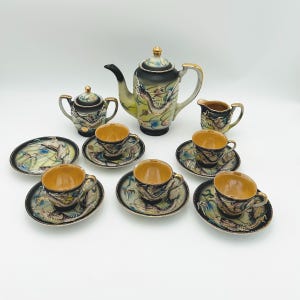 May include: A vintage tea set with a dragon motif. The set includes a teapot, sugar bowl, creamer, saucers, and teacups. The teacups and saucers are black with gold trim. The teapot is green and black with a gold handle.