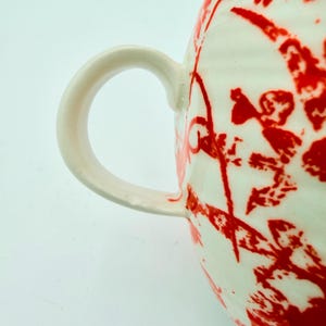 Może przedstawiać: Zbliżenie białego ceramicznego kubka z zakrzywionym uchwytem. Kubek ma odważny czerwony abstrakcyjny wz&oacute;r. Uchwyt jest w jednolitym białym kolorze. Kubek jest prawdopodobnie używany do picia napoj&oacute;w.