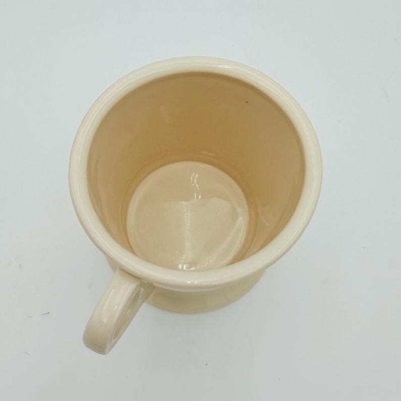 Puede incluir: Taza de cer&aacute;mica color crema con asa, vista desde arriba. La taza tiene una forma cil&iacute;ndrica ancha con una base redondeada y un asa peque&ntilde;a. El interior es de un color crema similar.