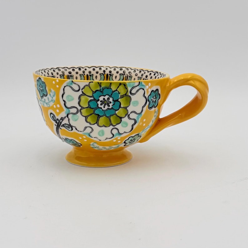 May include: A yellow ceramic teacup with a floral design. The cup features a large, stylized flower in shades of green and blue, set against a white background. The handle and base are also yellow, and the rim has a black and white dotted pattern.