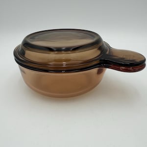 May include: A brown glass casserole dish with a matching lid and a handle. The dish is round with a slightly flared rim. The lid is also round and fits snugly on top. The handle is an extension of the dish and is easy to grip.