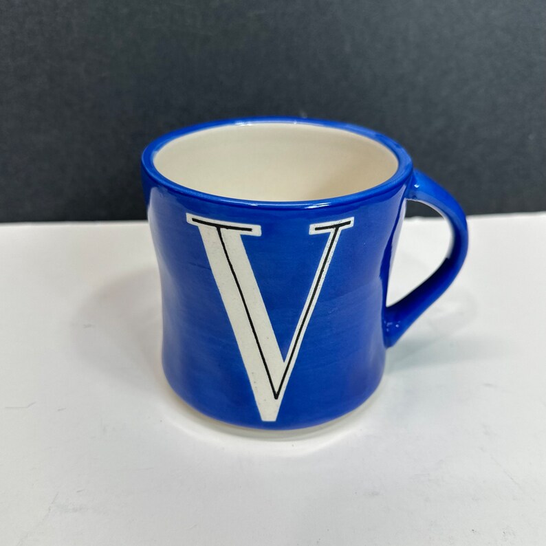 May include: A blue ceramic mug with a white interior and a curved handle. The exterior features a large white letter "V" with black trim. The mug is set against a white surface with a dark background.