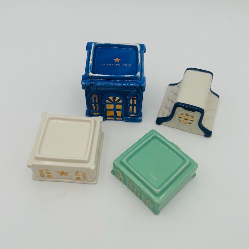 May include: Set of four miniature ceramic houses with gold accents. The houses are blue, white, green, and cream. The blue house has the text "Anthropologie" on the top.