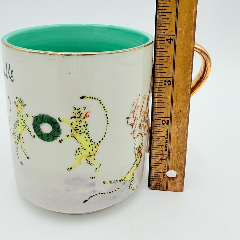 K&ouml;nnte beinhalten: Eine wei&szlig;e Keramiktasse mit goldenem Henkel und Rand, mit t&uuml;rkisfarbenem Innenraum. Die Tasse ist mit gelben Geparden mit schwarzen Flecken verziert, die einen gr&uuml;nen Kranz halten. Die Tasse ist etwa 10 cm hoch.