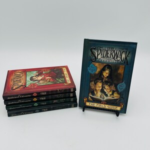 Puede incluir: Un conjunto de libros titulados "The Spiderwick Chronicles" y "The Field Guide". Los libros presentan portadas ilustradas y coloridas. "The Field Guide" se exhibe en un peque&ntilde;o soporte negro, mostrando su portada.