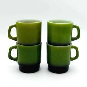 以下が含まれることがあります： ペアで積み重ねられた4つの緑色のガラスマグカップ。マグカップは丸みを帯びた上部と正方形のハンドルが付いています。マグカップの色は、上部の明るい緑色からベースの濃い緑色に変化します。