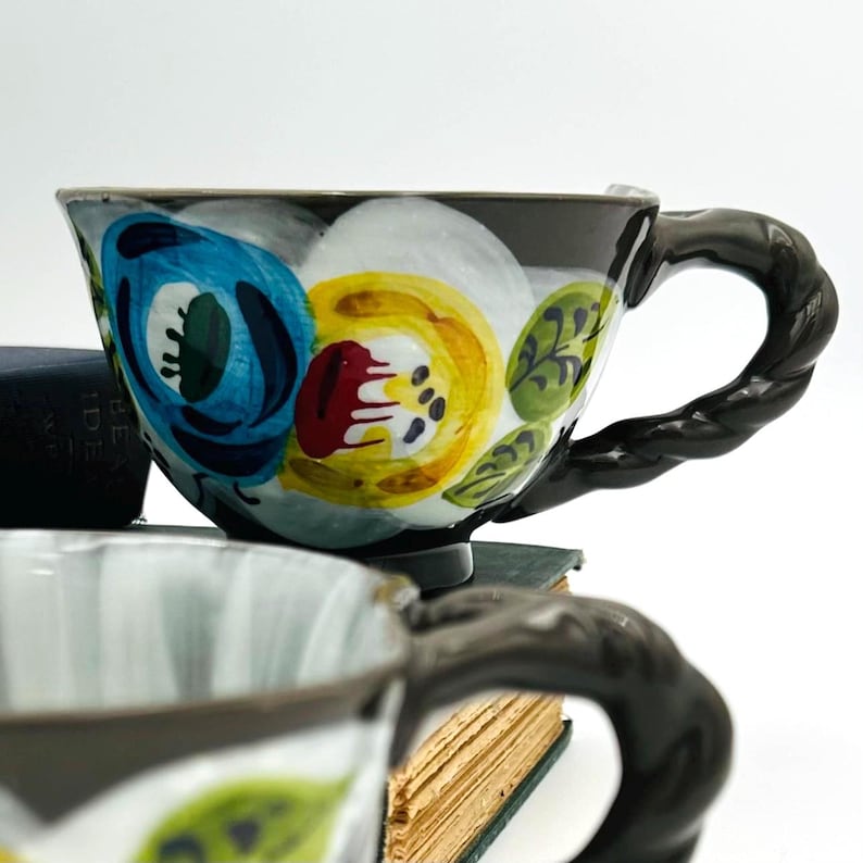 Puede incluir: Una taza de t&eacute; de cer&aacute;mica con asa y borde marr&oacute;n oscuro, con un dise&ntilde;o floral en azul, amarillo y rojo. La taza est&aacute; sobre una pila de libros, con otra taza en primer plano.