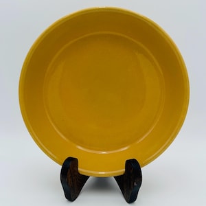 Puede incluir: Un plato redondo de cerámica amarillo mostaza con un borde ligeramente elevado. El plato se exhibe sobre un soporte de madera oscura. La superficie del plato tiene una apariencia sutil y texturizada. Es un artículo de cocina.