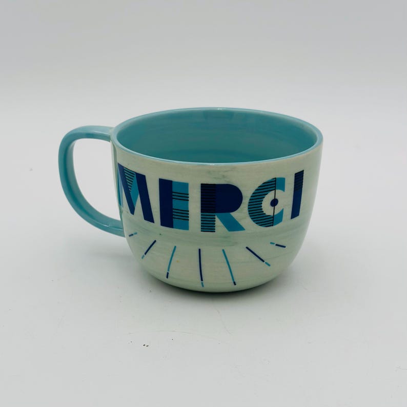 May include: A light blue ceramic mug with a white interior and a blue and white geometric design that says "MERCI".