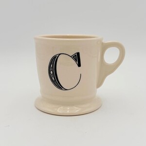 Puede incluir: Taza de cer&aacute;mica color crema con la letra negra "C" en la parte delantera. La taza tiene un asa y una base de pedestal. La letra "C" est&aacute; en una fuente decorativa con un dise&ntilde;o punteado.