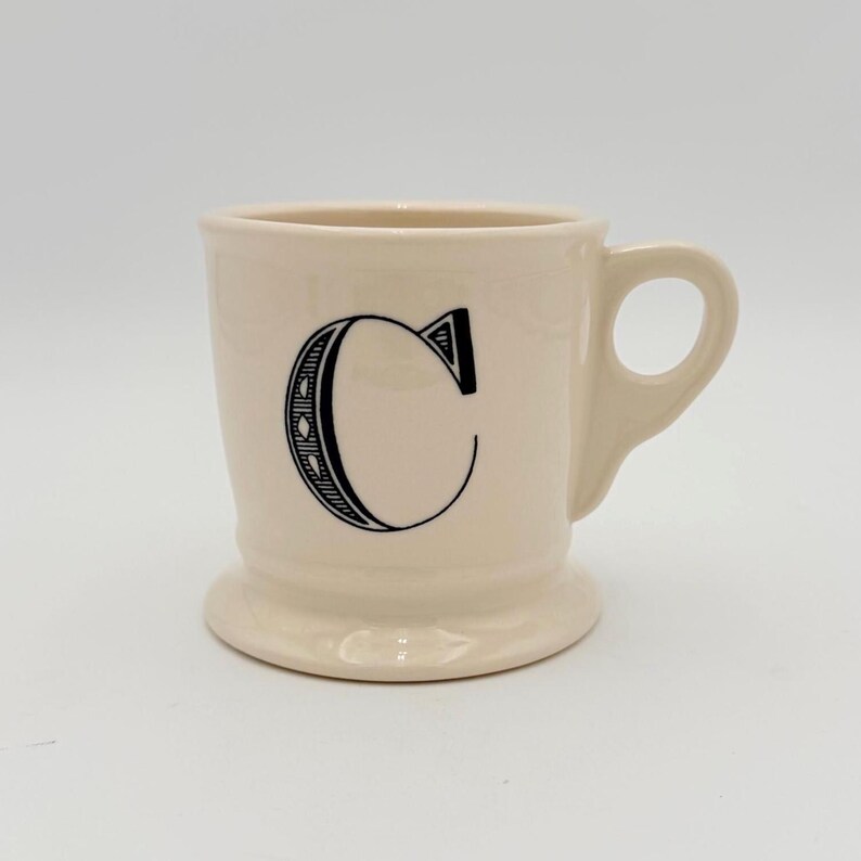 Puede incluir: Taza de cer&aacute;mica color crema con la letra negra "C" en la parte delantera. La taza tiene un asa y una base de pedestal. La letra "C" est&aacute; en una fuente decorativa con un dise&ntilde;o punteado.