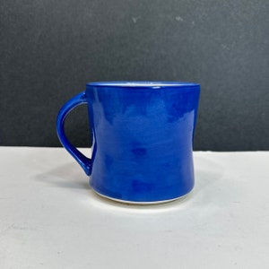 May include: A cobalt blue ceramic mug with a curved handle. The mug has a glossy finish and a white interior. The mug is sitting on a white surface with a black background.