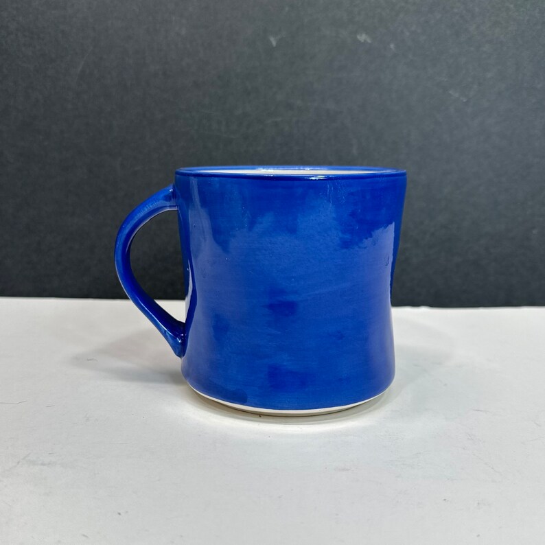May include: A cobalt blue ceramic mug with a curved handle. The mug has a glossy finish and a white interior. The mug is sitting on a white surface with a black background.