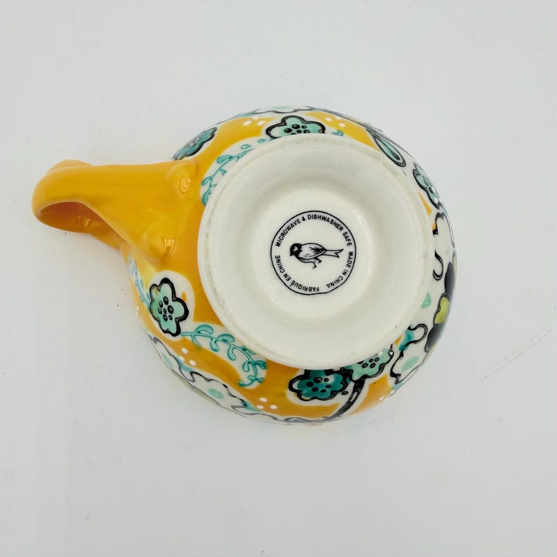 May include: Yellow ceramic spoon rest with a handle. The spoon rest has a floral pattern in teal, black, and white. The bottom of the spoon rest has the text "Microwave & Dishwasher Safe" and "Made in China".