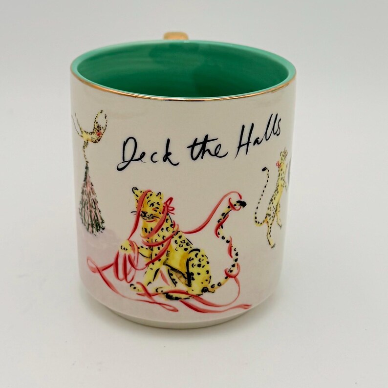 K&ouml;nnte beinhalten: Cremefarbene Keramiktasse mit gr&uuml;nem Innenraum und Goldrand. Die Tasse zeigt ein handgezeichnetes Design eines gelben Leoparden mit roter Schleife und den Worten "Deck the Halls". Eine festliche Tasse.