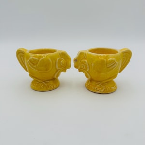 May include: Two yellow ceramic chicken egg cups. Each cup has a white design and the words "Made in USA" on the bottom.