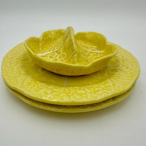 May include: A set of three yellow ceramic plates and a small bowl. The plates and bowl are shaped like cabbage leaves with white vein detailing. The bowl has a handle. The set is a bright, cheerful addition to any kitchen.