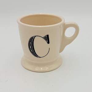 Puede incluir: Taza de cer&aacute;mica color crema con la letra "C" negra en la parte delantera. La taza tiene una base redondeada, un asa y un acabado liso y brillante. El interior es de un color crema m&aacute;s claro.