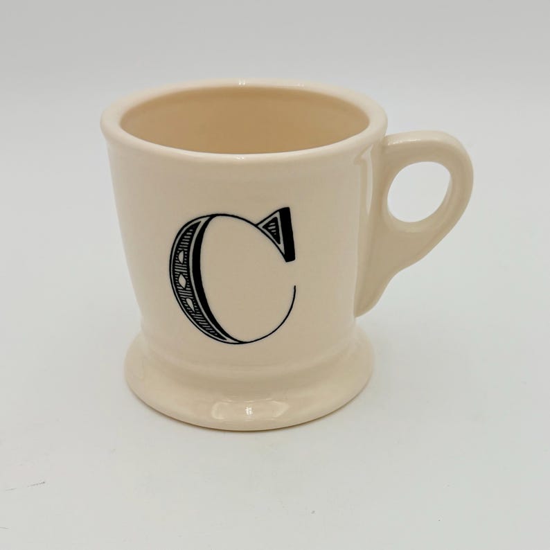 Puede incluir: Taza de cer&aacute;mica color crema con la letra "C" negra en la parte delantera. La taza tiene una base redondeada, un asa y un acabado liso y brillante. El interior es de un color crema m&aacute;s claro.