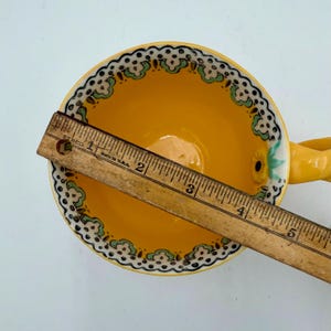 May include: A yellow teacup with a decorative handle and a floral design. The rim features a white and black pattern with green accents. A wooden ruler is placed across the cup, measuring approximately 5 inches.