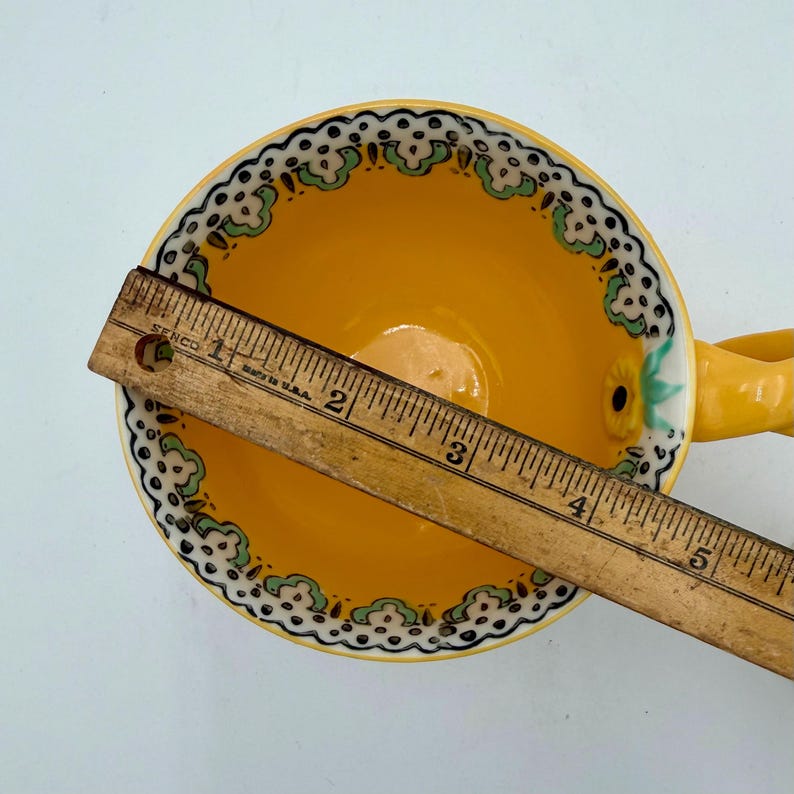 May include: A yellow teacup with a decorative handle and a floral design. The rim features a white and black pattern with green accents. A wooden ruler is placed across the cup, measuring approximately 5 inches.