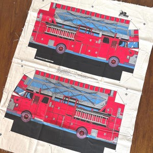 May include: A white fabric panel featuring two printed red fire engine designs with blue ladders, black wheels, and F.D. lettering. The fabric includes sewing instructions, registration marks, and the text The Toy Works, New York 1988.