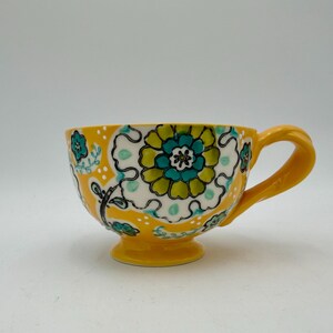May include: A yellow ceramic teacup with a large handle and a pedestal base. The cup is decorated with a floral pattern in shades of green, teal, and white against a yellow background. The design includes a large central flower and smaller floral accents.