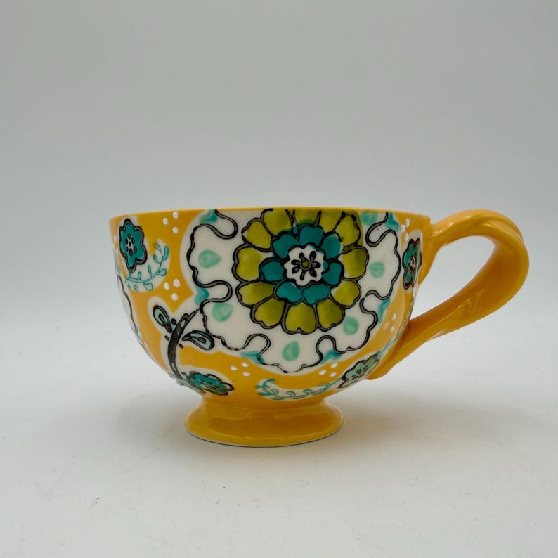 May include: A yellow ceramic teacup with a large handle and a pedestal base. The cup is decorated with a floral pattern in shades of green, teal, and white against a yellow background. The design includes a large central flower and smaller floral accents.