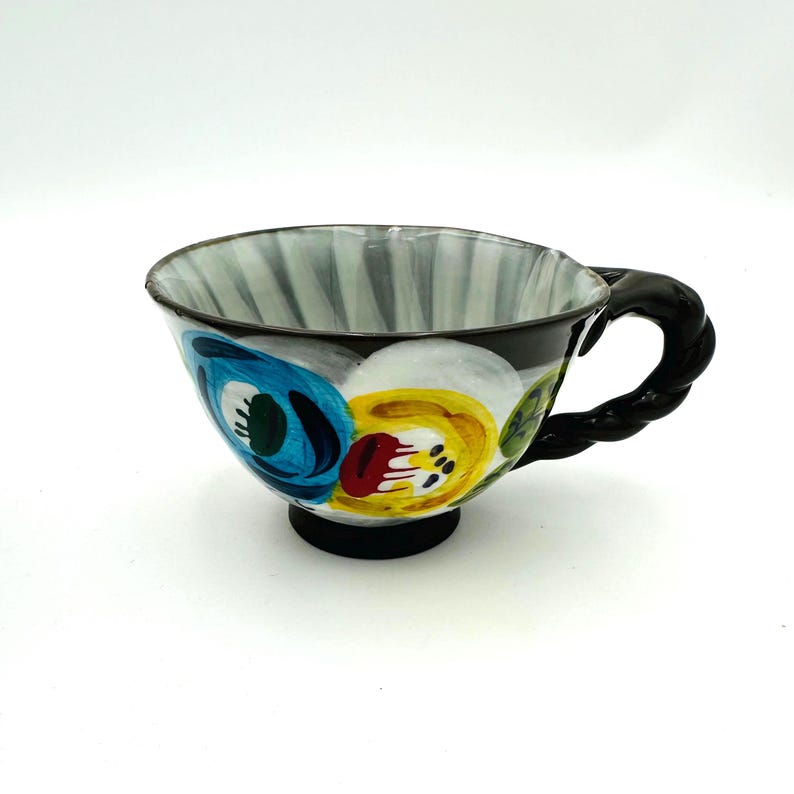 Puede incluir: Una taza de t&eacute; de cer&aacute;mica pintada a mano con asa y base negras. La taza presenta un dise&ntilde;o floral en azul, amarillo y rojo sobre un fondo negro. El interior tiene rayas verticales grises y blancas.
