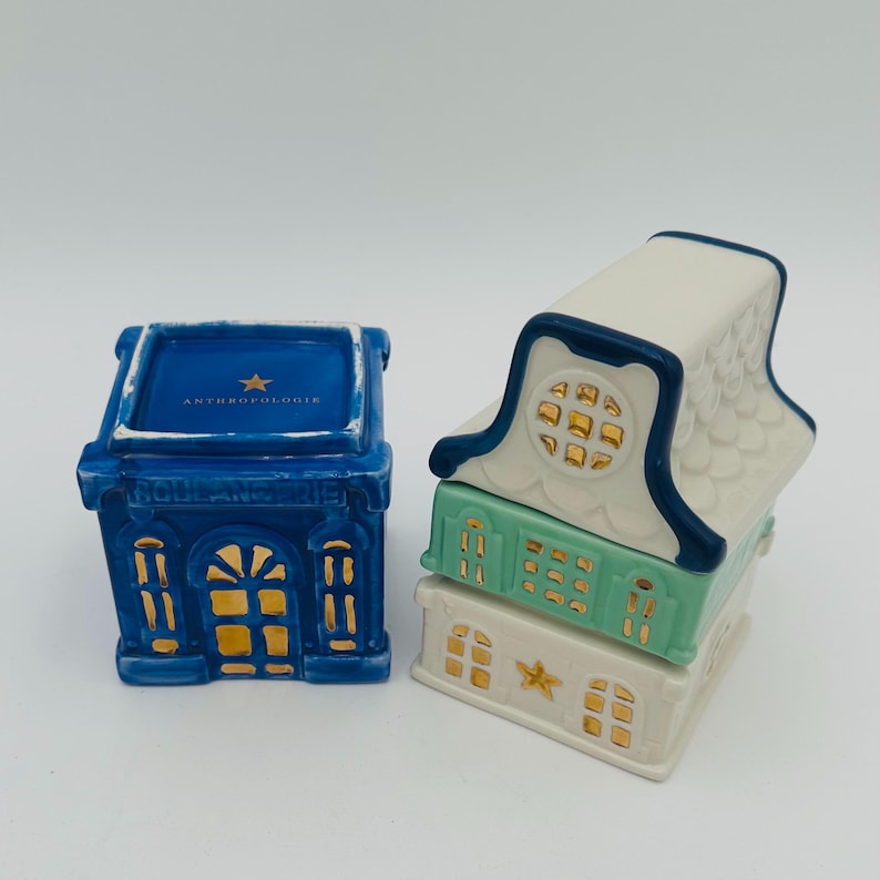 May include: Two ceramic houses with gold accents. The larger house is blue with white trim and the text "Anthropologie" on the top. The smaller house is white with blue trim and a green middle section. Both houses have gold stars on the front.