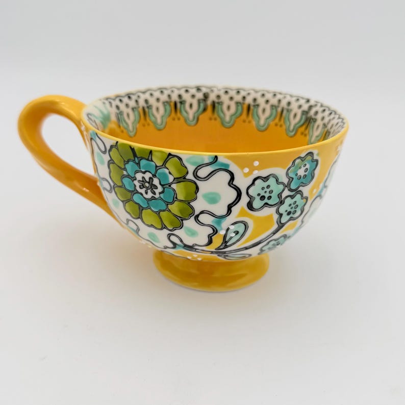 May include: A yellow ceramic teacup with a floral design in shades of blue, green, and white. The cup has a rounded base, a curved handle, and a decorative rim. The interior of the cup is yellow.