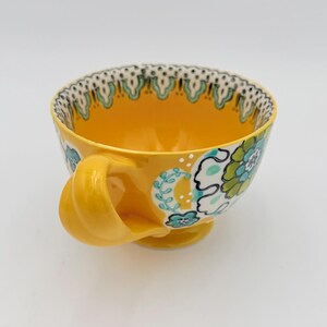 May include: Yellow ceramic teacup with a floral design in teal, white, and green. The cup has a rounded handle and a decorative rim with a teal and black pattern. The cup sits on a small foot.