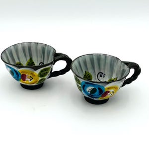 Puede incluir: Dos tazas de t&eacute; de cer&aacute;mica con asas y bases negras. Las tazas tienen un interior a rayas grises y blancas y est&aacute;n decoradas con coloridos dise&ntilde;os florales en azul, amarillo y rojo. Las tazas est&aacute;n sobre un fondo blanco.