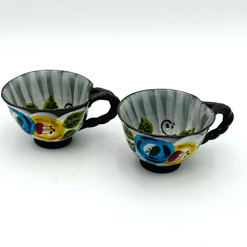 Puede incluir: Dos tazas de t&eacute; de cer&aacute;mica con asas y bases negras. Las tazas tienen un interior a rayas grises y blancas y est&aacute;n decoradas con coloridos dise&ntilde;os florales en azul, amarillo y rojo. Las tazas est&aacute;n sobre un fondo blanco.