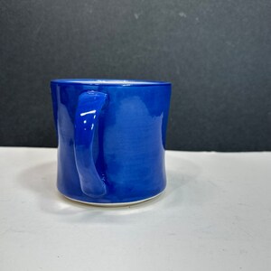 May include: A cobalt blue ceramic mug with a curved handle. The mug has a glossy finish and a white base. The mug is sitting on a white surface with a black background.