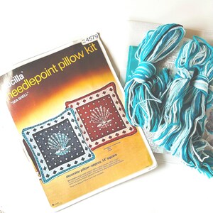 May include: A vintage Lucilla needlepoint pillow kit labeled "SEA SHELL." The kit includes a pattern for a decorator pillow, approximately 14 inches square, with a seashell design. Bundles of blue and white yarn are also visible.