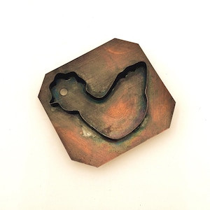 May include: A copper-colored metal cookie cutter in the shape of a chicken. The cutter has a rustic, aged look and is slightly tarnished.