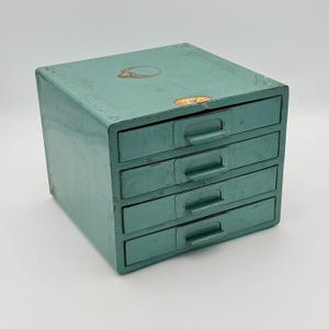 May include: A vintage teal metal storage cabinet with four drawers. The cabinet has a rectangular shape with rounded corners and small metal handles on each drawer. There are two stickers on the top of the cabinet.
