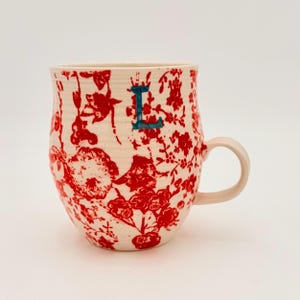 Może przedstawiać: Kremowy ceramiczny kubek z czerwonym kwiatowym wzorem. Kubek ma jasnor&oacute;żowy uchwyt i turkusową literę "L". Wz&oacute;r to powtarzający się motyw kwiat&oacute;w i liści. Unikalny element wyposażenia kuchni.