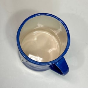 May include: A blue ceramic mug with a white interior. The mug has a blue rim and a matching blue handle. The mug is empty and sits on a white surface. The mug is viewed from above.