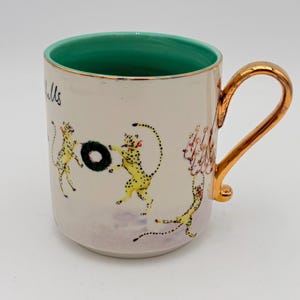 K&ouml;nnte beinhalten: Cremefarbene Keramiktasse mit goldenem Henkel und Rand. Der Innenraum ist gr&uuml;n. Die Tasse zeigt Illustrationen von gelben Geparden mit schwarzen Flecken, die mit einem gr&uuml;nen Kranz spielen. Die Tasse hat den Text "Hills" auf der Seite.